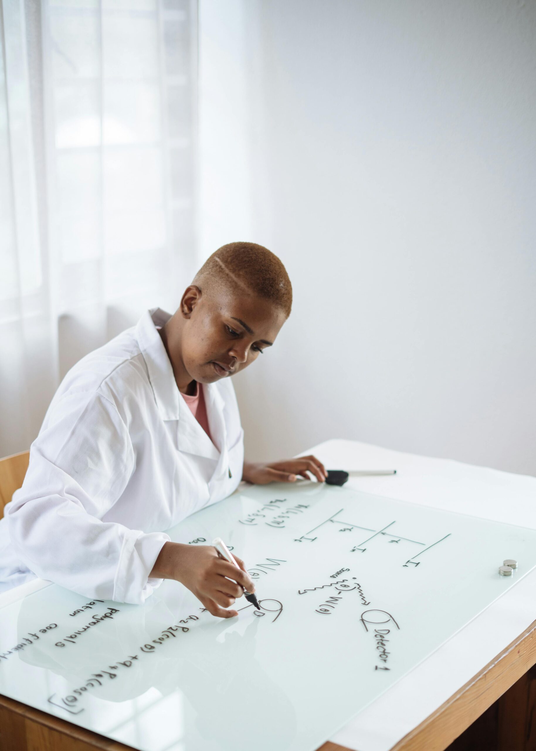 African-American female scientist in lab coat writing scientific equations on a glass board.